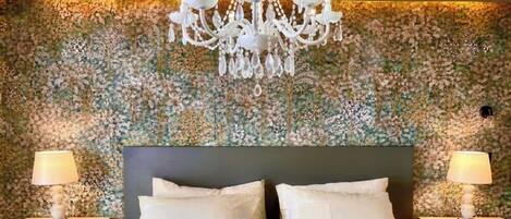 Quarto com papel de parede floral, um lustre, duas mesas de cabeceira com luminárias e uma cama com almofadas.