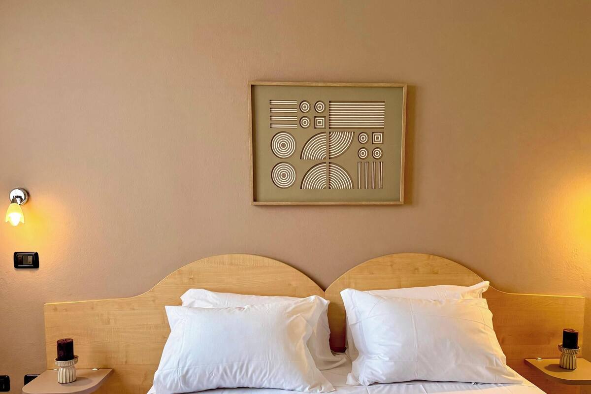 Habitación de hotel con cabecera de madera, dos almohadas blancas, una obra de arte colgada en la pared y dos lámparas de noche.