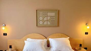 Een hotelkamer met een houten hoofdeinde, twee witte kussens, een aan de muur bevestigd kunstwerk en twee bedlampjes.