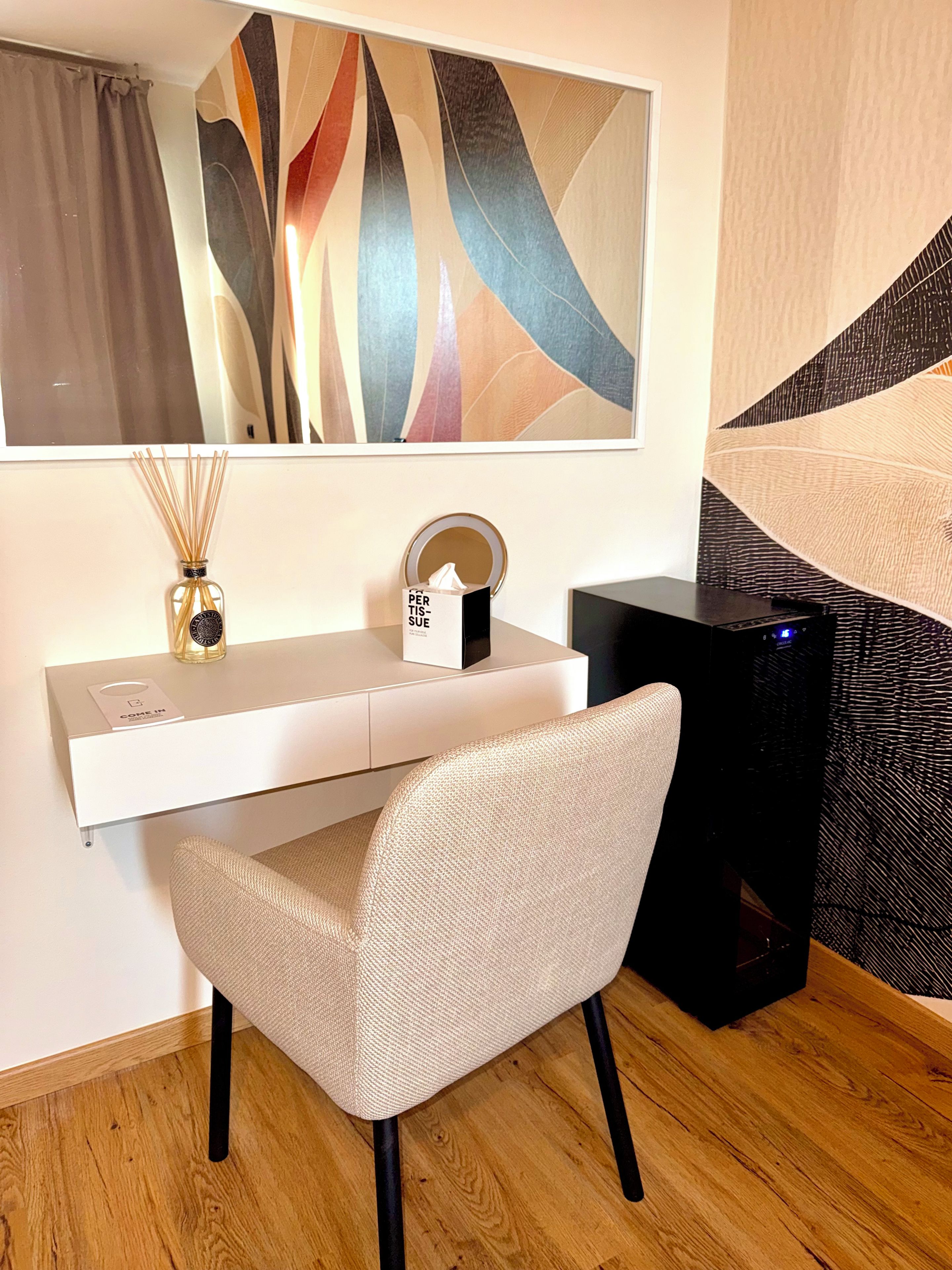 Une chambre moderne avec un bureau blanc, une chaise, un grand tableau abstrait, un miroir, une boîte de mouchoirs et un haut-parleur noir.