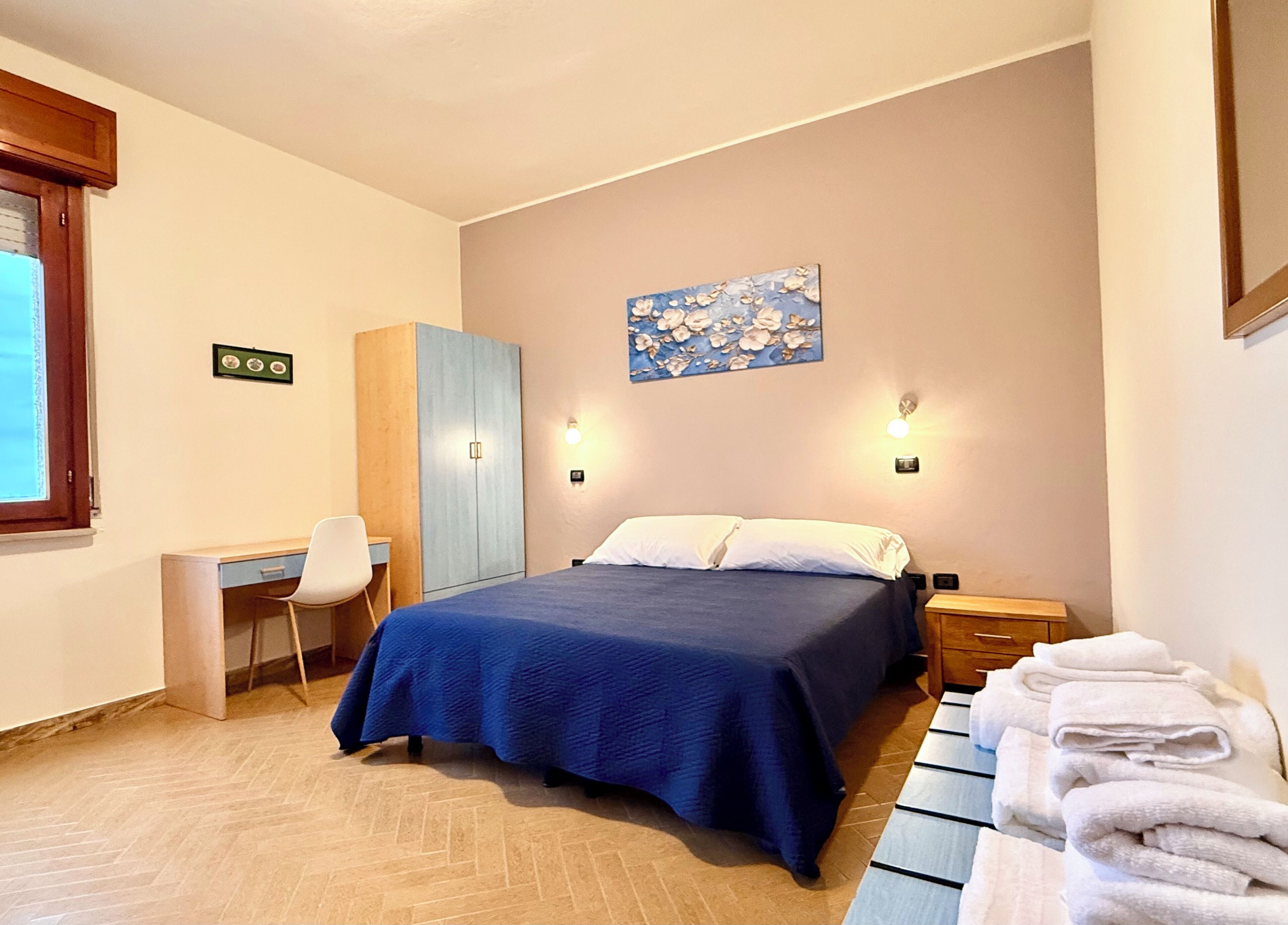 A bedroom with a bed, a desk, a chair, a wardrobe, and a wall-mounted light fixture.