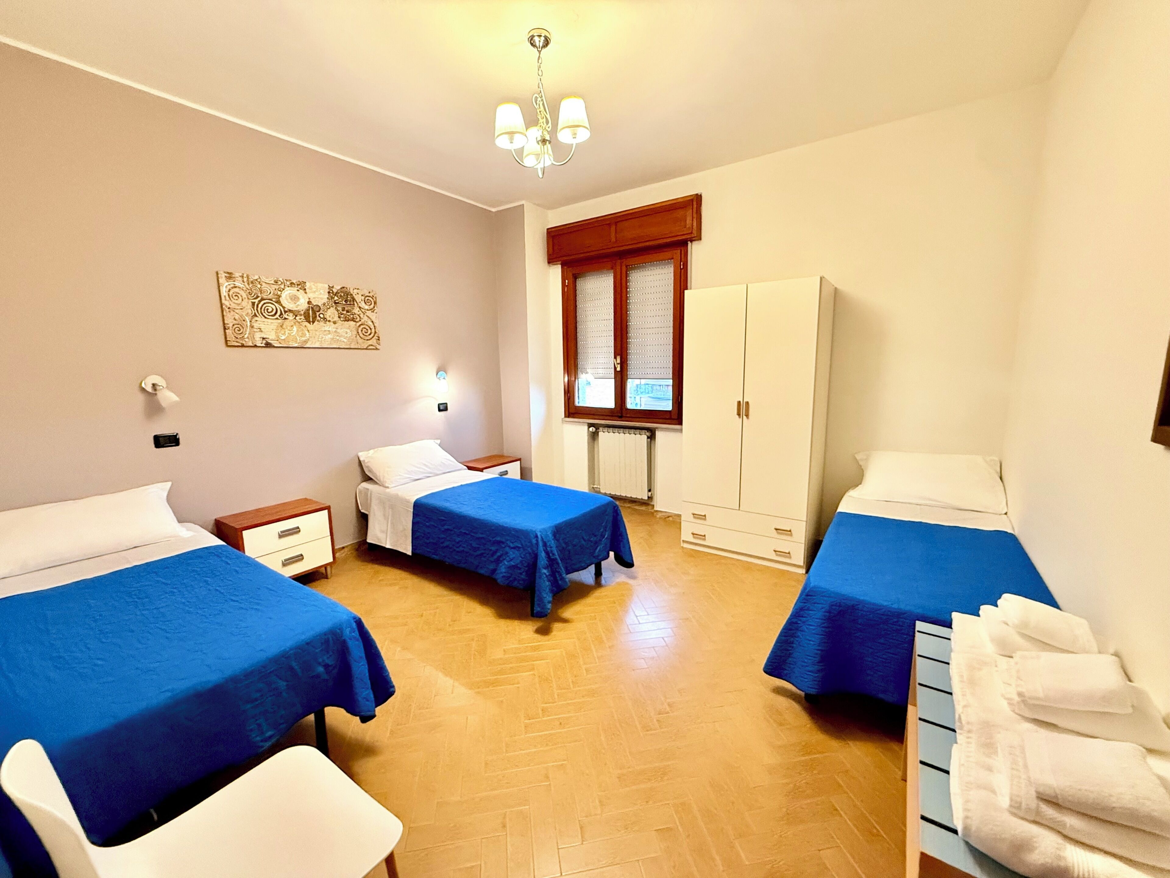 A hotel room with two beds, a wardrobe, a window with blinds, a chandelier, and a wall-mounted artwork.