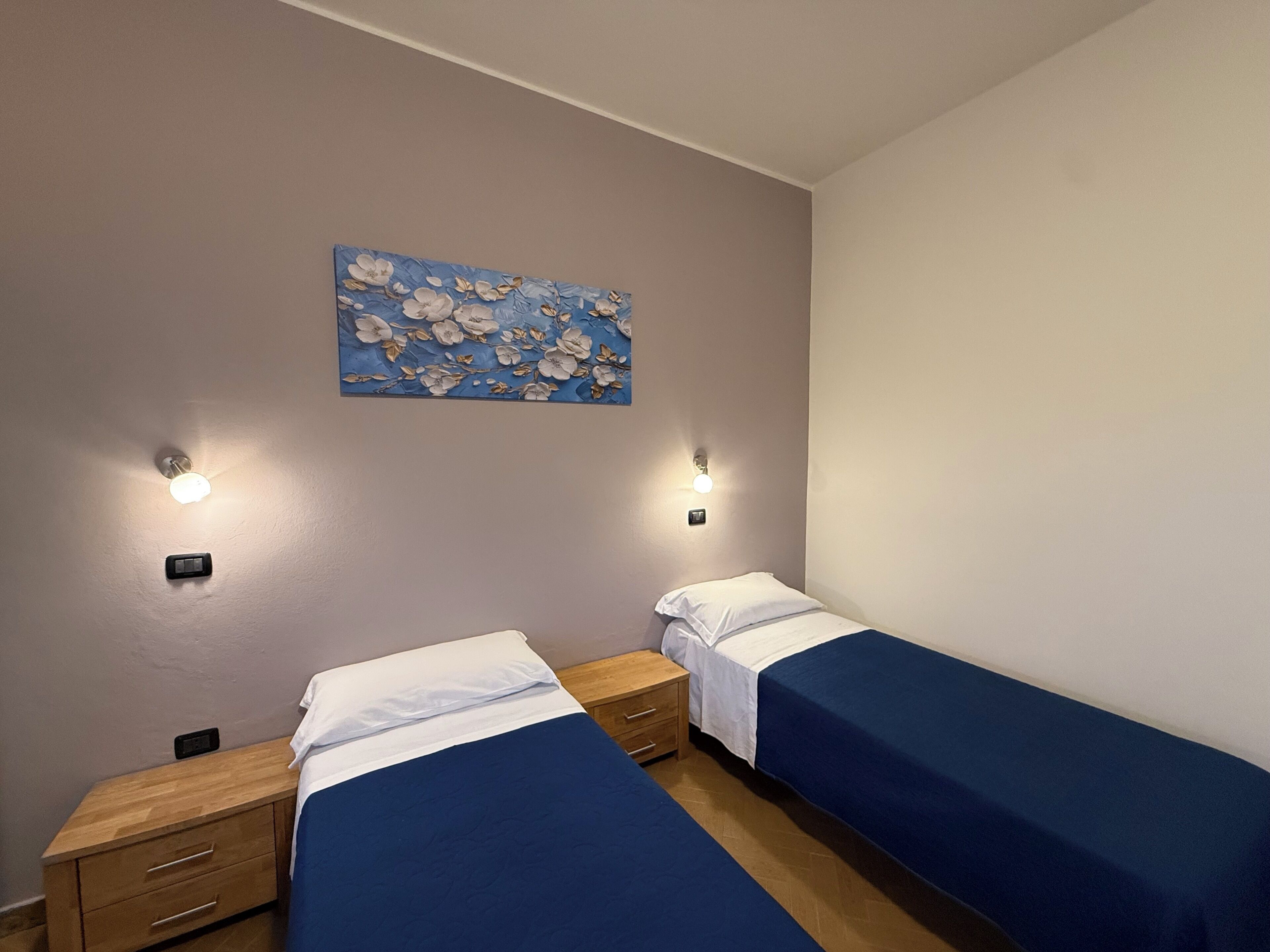 Un dormitorio con dos camas individuales, una mesita de noche con cajones, un aplique de pared y un cuadro con motivos florales.