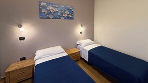 A bedroom with two single beds, a nightstand with drawers, a wall-mounted light fixture, and a floral painting. - HOTEL IL MULINO (Casalgrande)