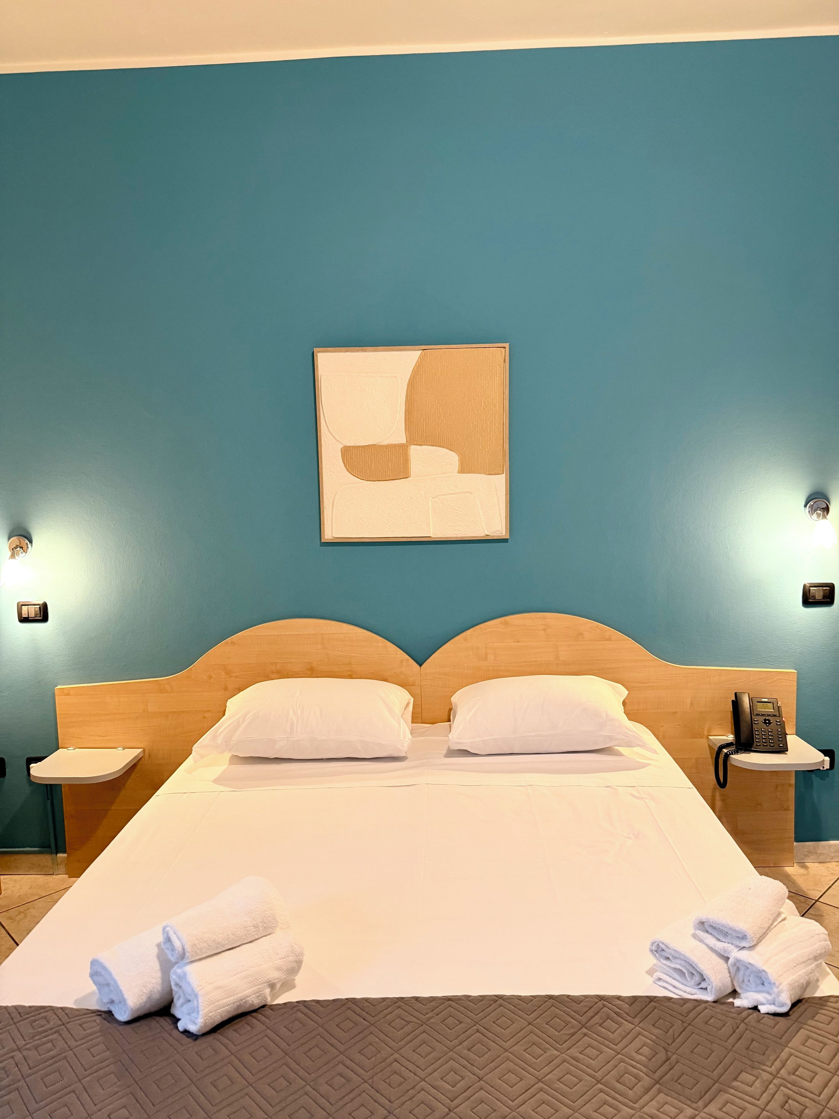 Una habitación de hotel con una cama, dos mesitas de noche, un teléfono en la pared y un cuadro.