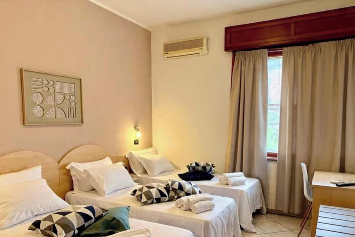Habitación de hotel con dos camas, un escritorio, una silla, ventana con cortinas, unidad de aire acondicionado y una lámpara de pared.