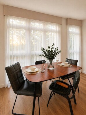 A dining room with a wooden table, black leather chairs, a vase with green foliage, and a window with sheer curtains. - Stylish 2-Bed 2-Bath Apartment with Parking, Lift & Balcony - London & Airports (Croydon)