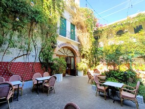 Eine Terrasse im Freien mit Korbmöbeln, ein Gebäudeeingang mit einem Schild und Grünpflanzen.