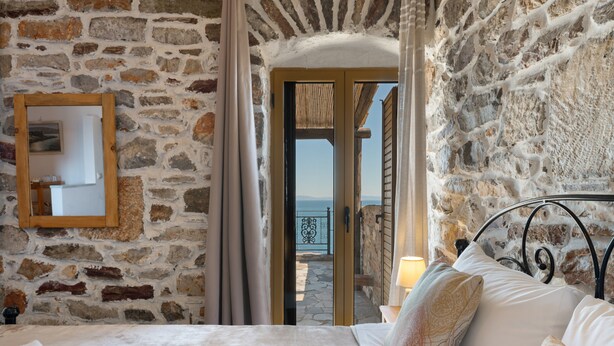 Una camera da letto con pareti in pietra, un letto con lenzuola bianche, uno specchio con cornice in legno e vista su un balcone attraverso una porta aperta.