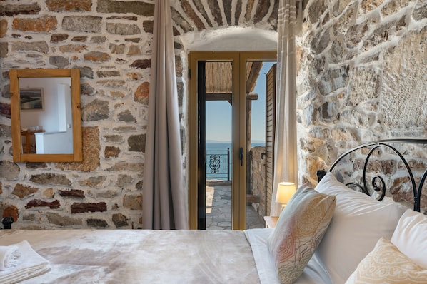 Ein Schlafzimmer mit Steinwänden, einem Bett mit weißen Leinen, einem spiegelnden Rahmen aus Holz und Blick durch eine offene Tür auf einen Balkon.