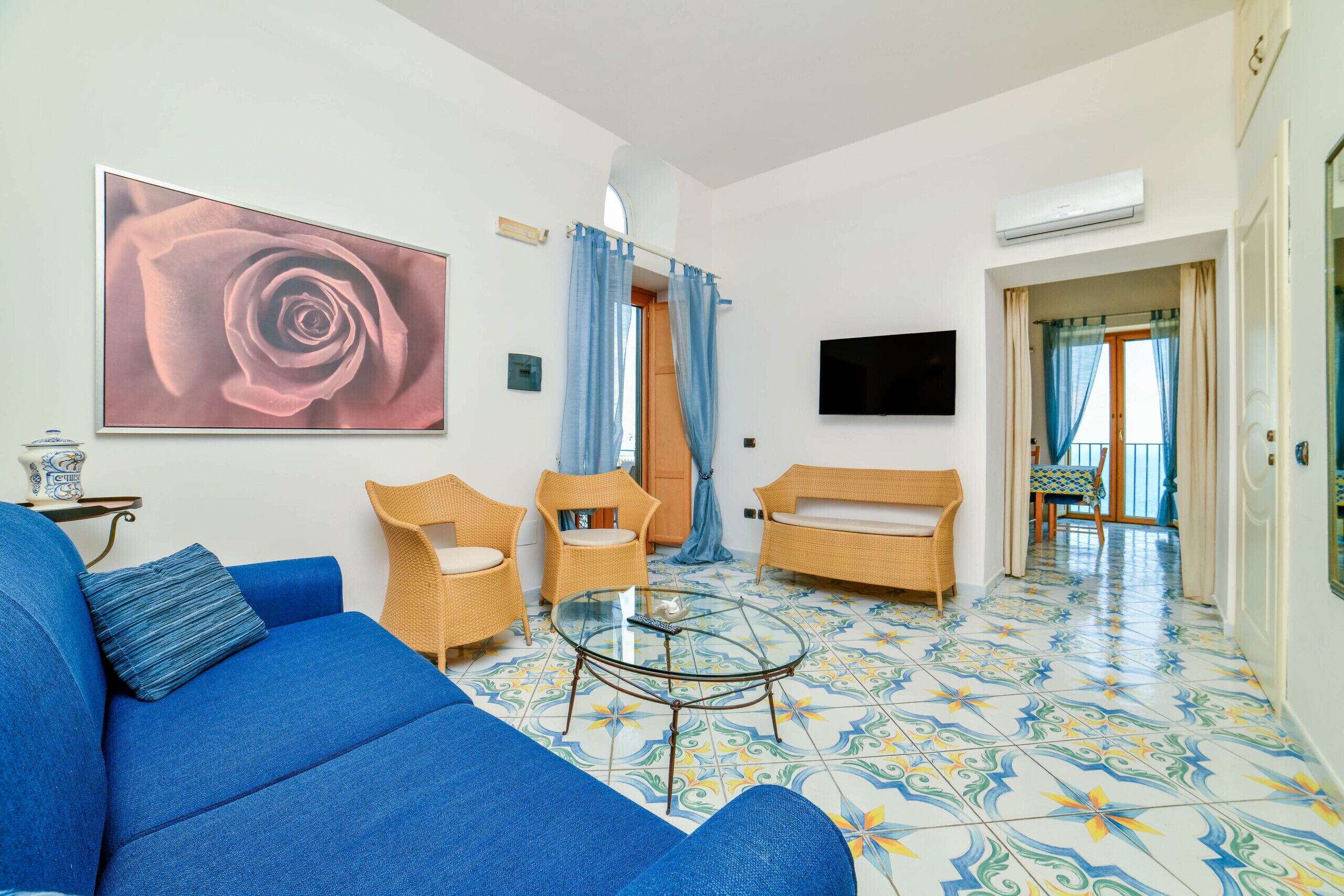 A living room with a blue sofa, two wicker chairs, a glass coffee table, a large rose painting, and a TV mounted on the wall.