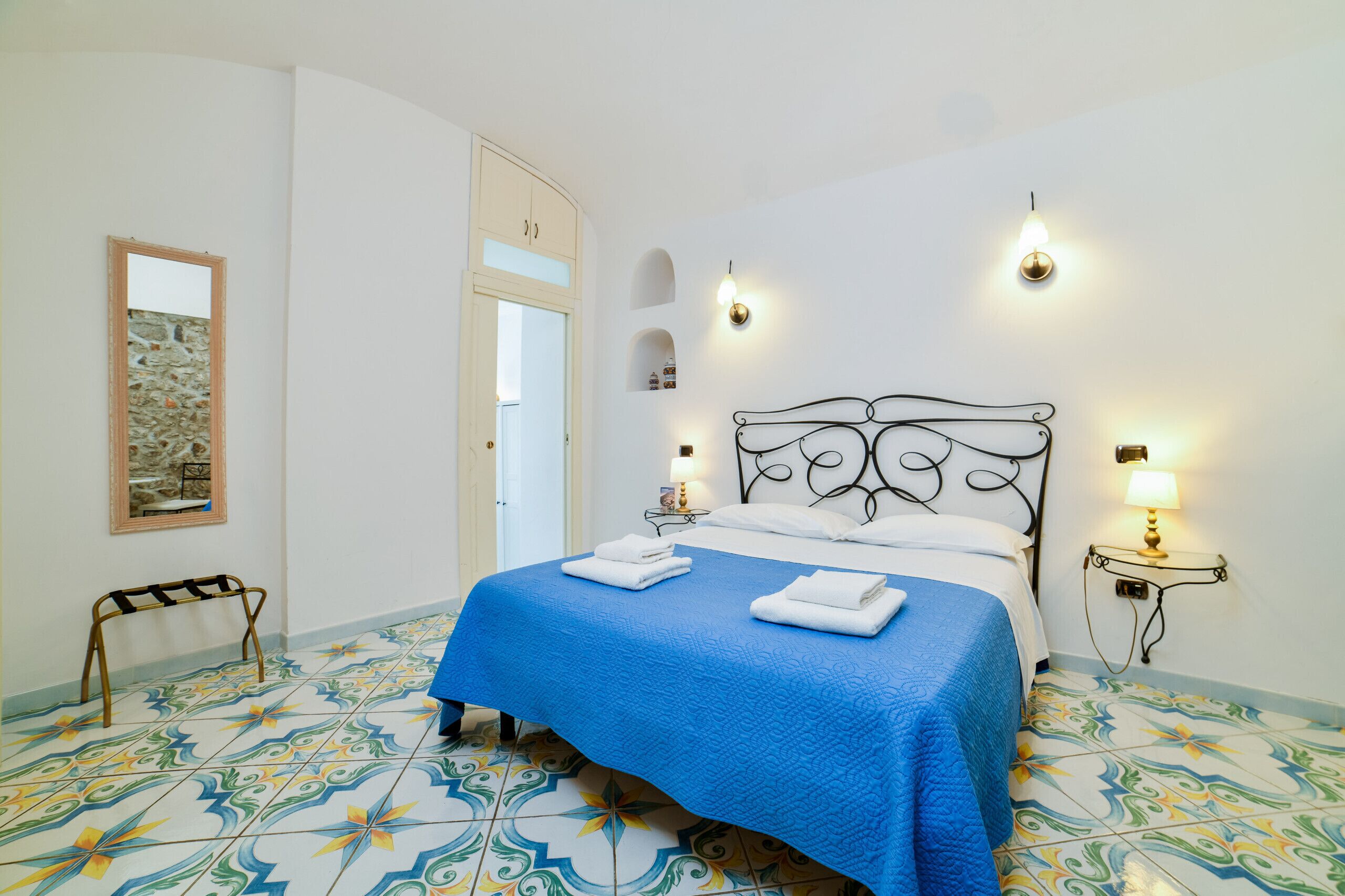 A bedroom with a bed, a nightstand, a wall-mounted lamp, a mirror, and a door.