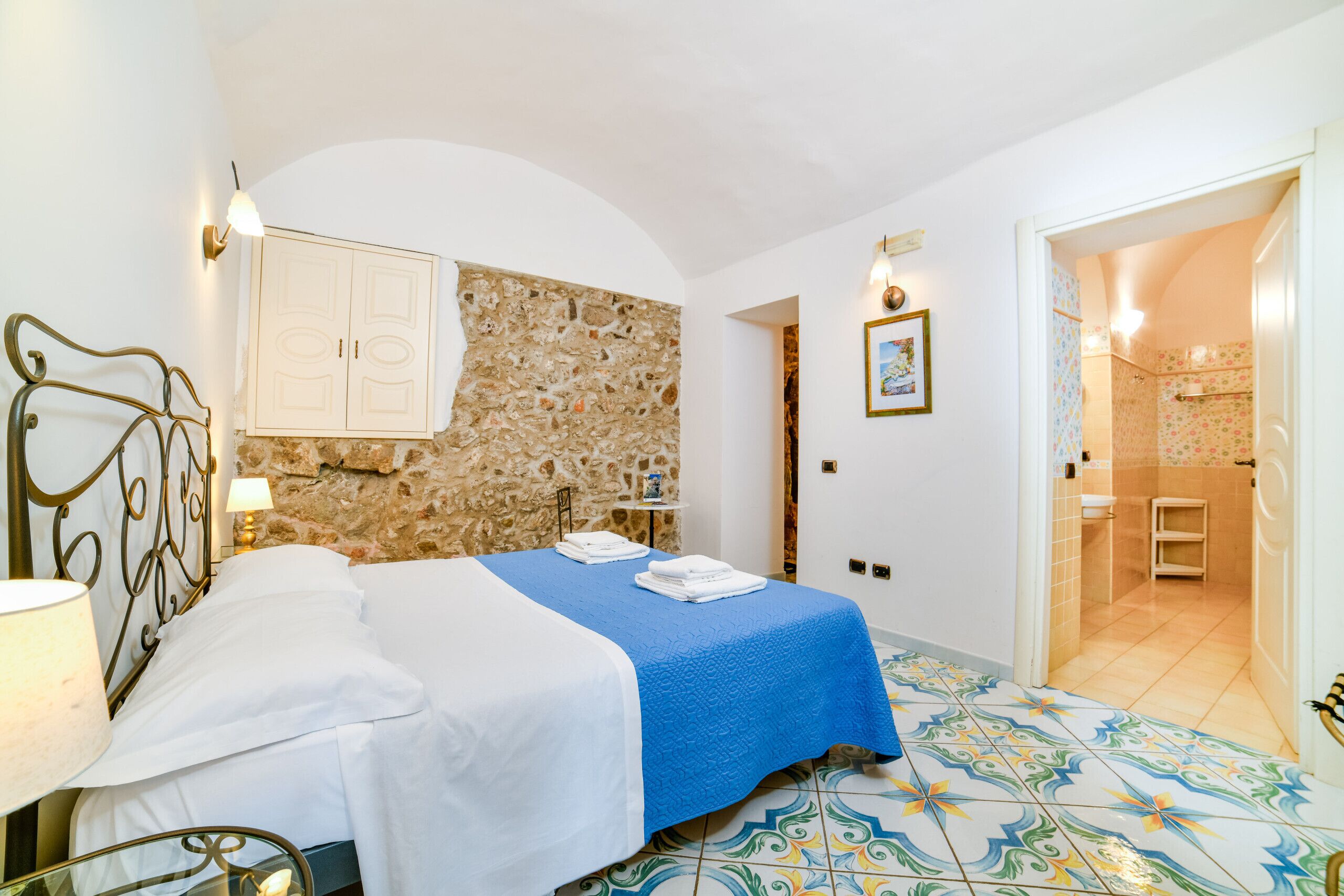 Een slaapkamer met een stenen muur, een bed met witte lakens, een blauwe spreideken en een betegelde vloer.