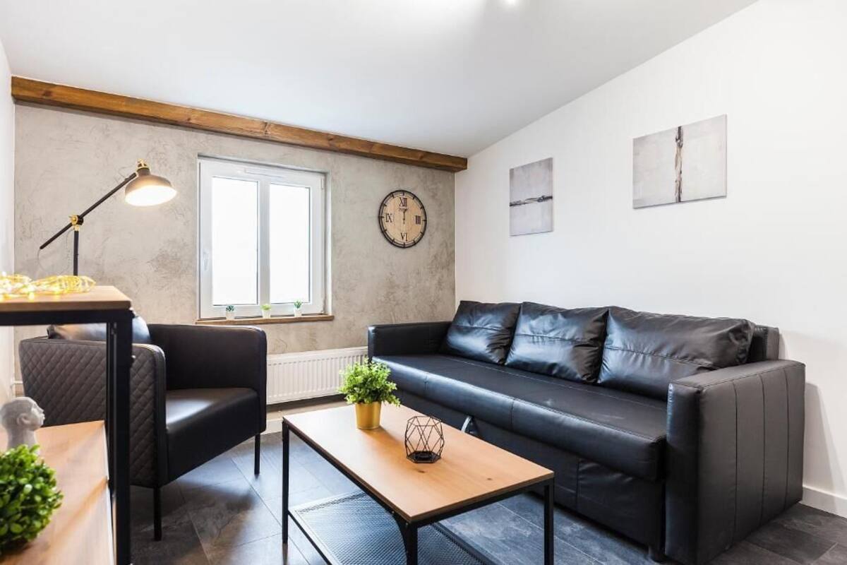 A modern living room with a black leather sofa, a wooden coffee table, a black armchair, a floor lamp, and a wall clock.
