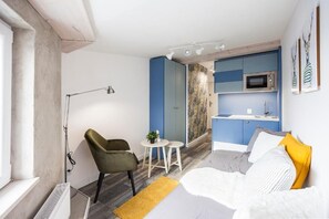 A compact room with a bed, a desk, a chair, a small table with a plant, and a kitchenette. - Book-Apart Fabryczna (Łódź)