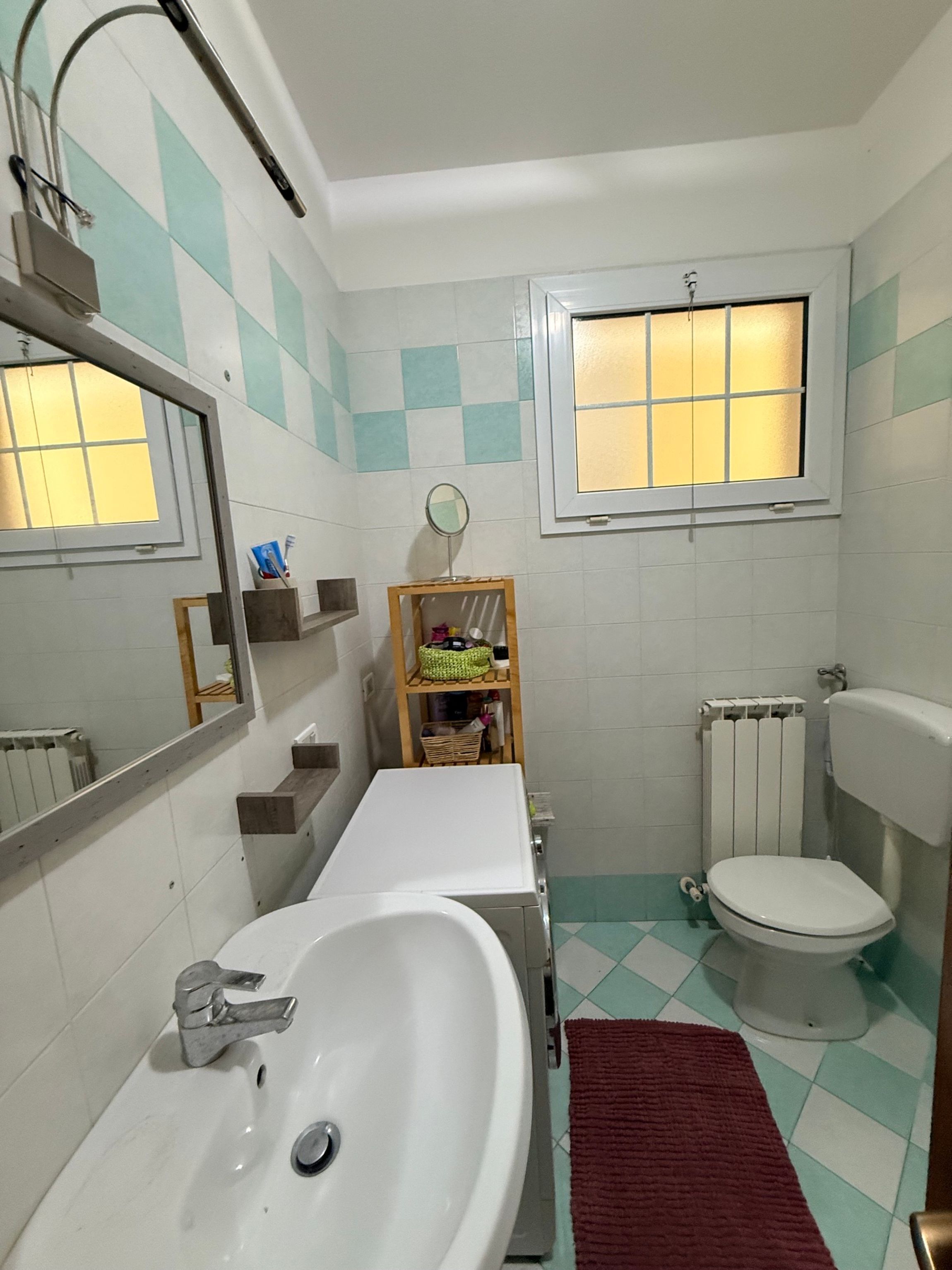A bathroom with a white sink, a mirror, a toilet, a window, and a radiator.