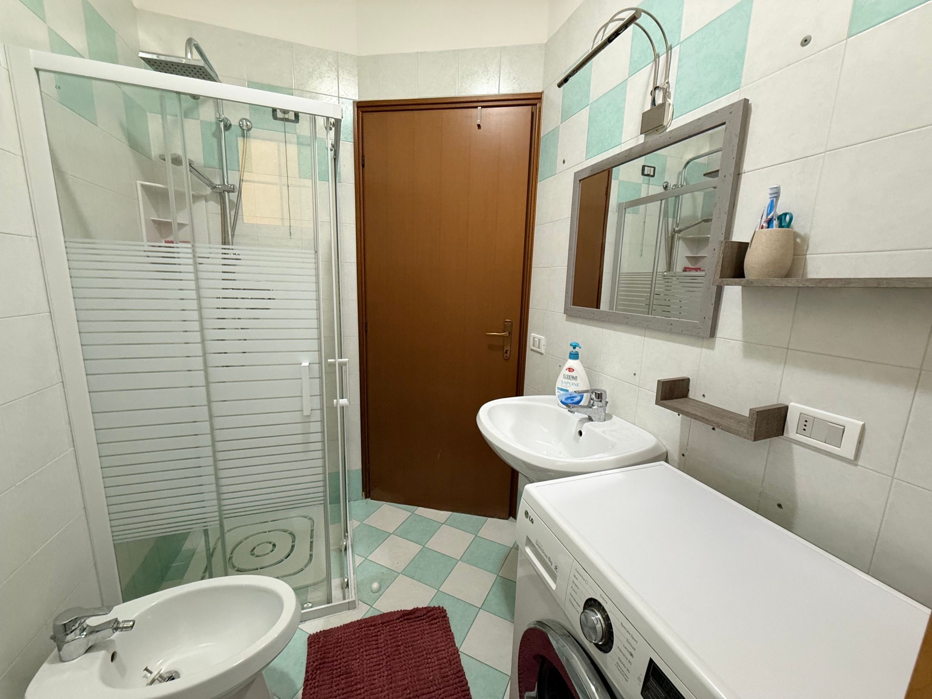A bathroom with a glass shower enclosure, a sink, a mirror, and a washing machine.