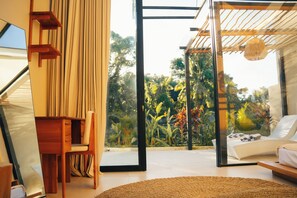 Ein modernes Zimmer mit Schreibtisch aus Holz, einem Sessel, einem Bett mit weißer Bettwäsche und Blick auf üppiges Grün durch große Glaswände.