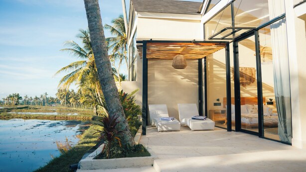 Ett modernt hus med en inglasad veranda, två solstolar och utsikt över en vattenyta och palmer.