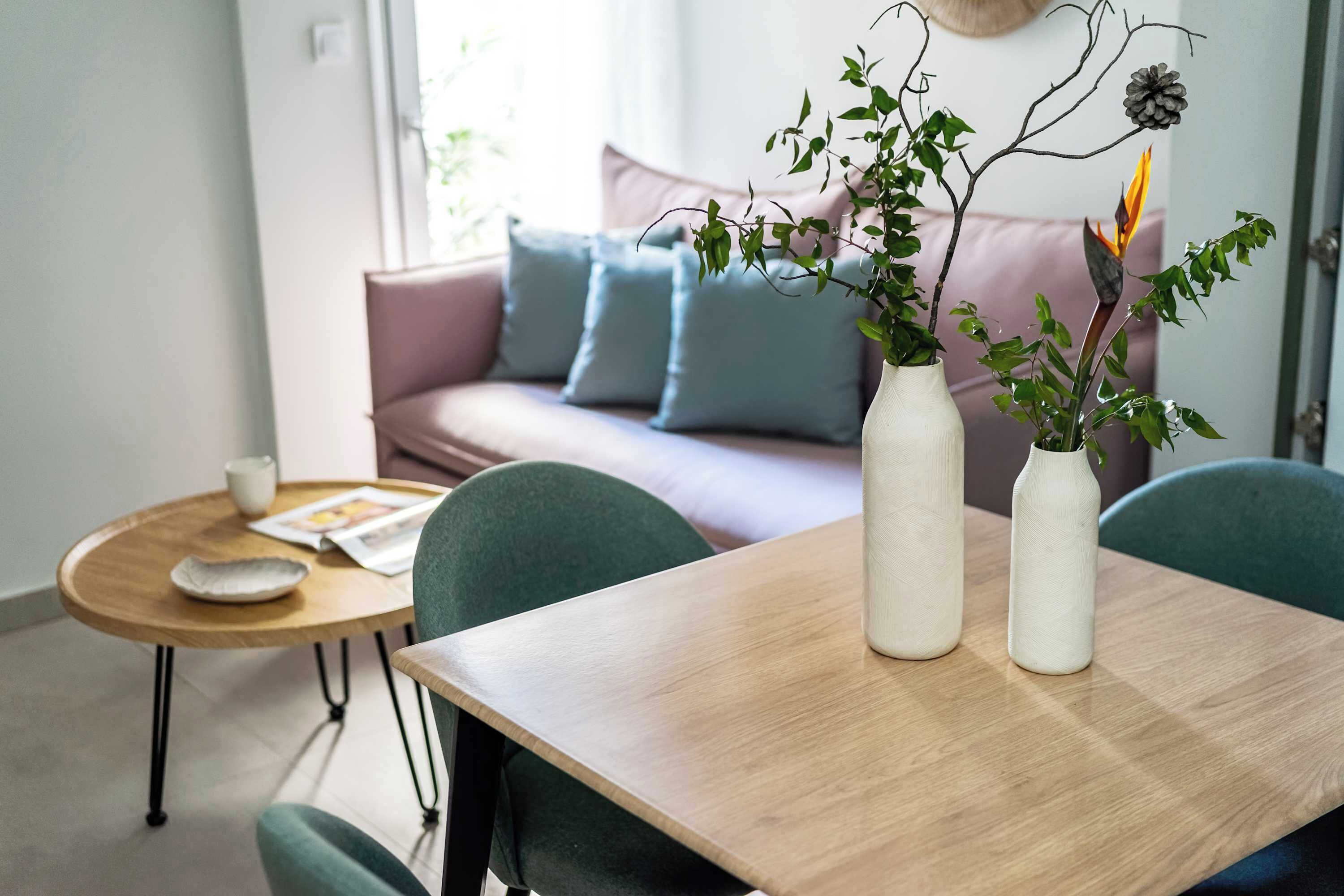 A modern living room with a wooden table, two white vases with plants, a green chair, a pink sofa with blue cushions, and a wooden coffee table with a magazine and a cup.