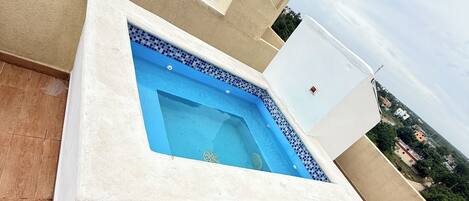 Una piscina en la azotea con borde de azulejos, rodeada por un edificio color crema con una ventana y una puerta.