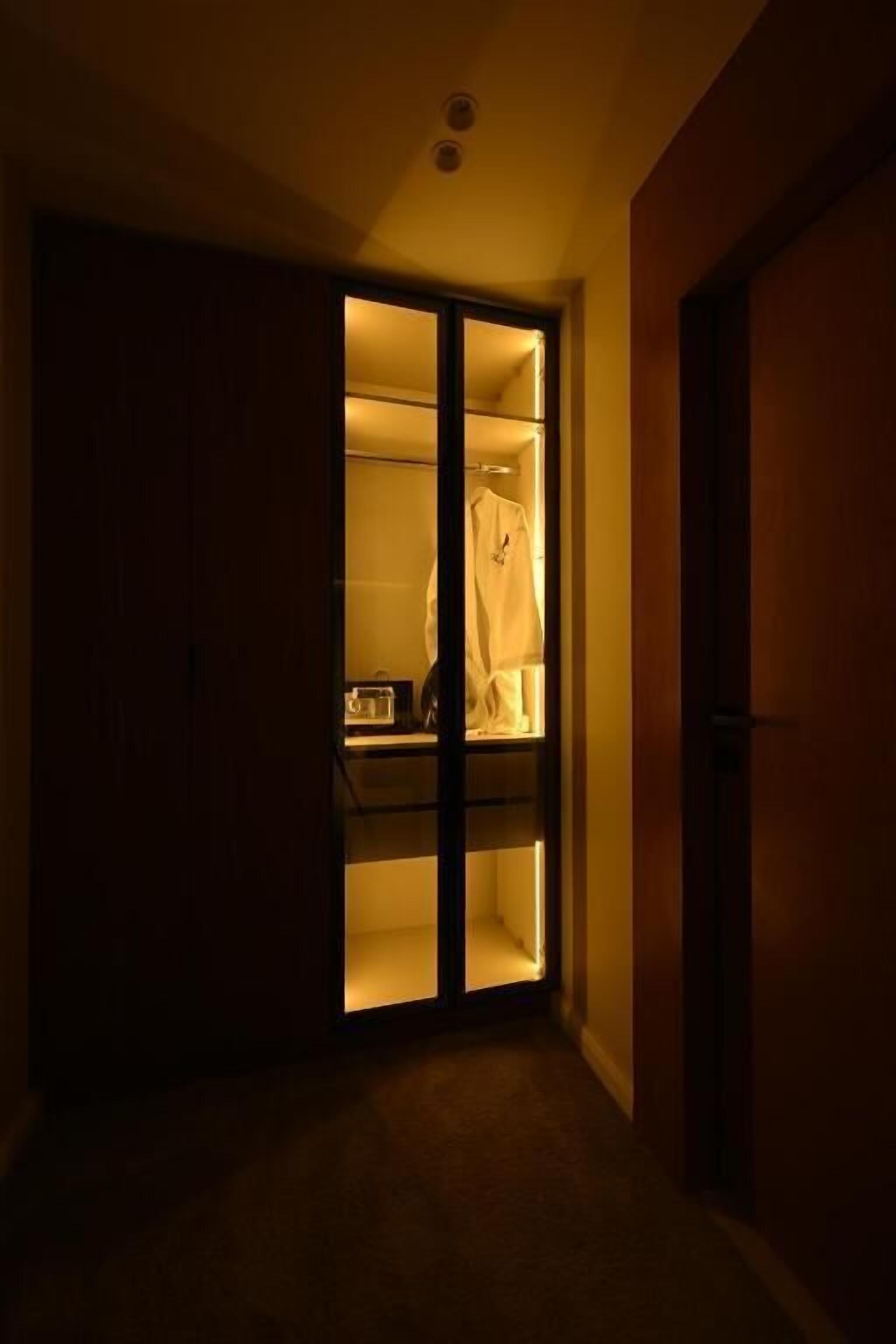 Un couloir faiblement éclairé, avec une porte coulissante donnant sur une pièce où se trouvent un lit et une table de chevet.