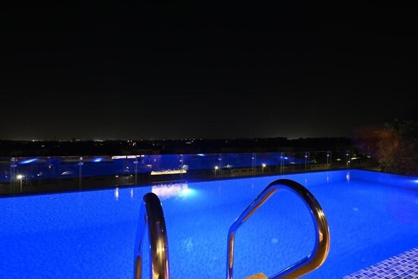 Vista nocturna de una piscina con una escalera metálica y el horizonte de la ciudad al fondo.