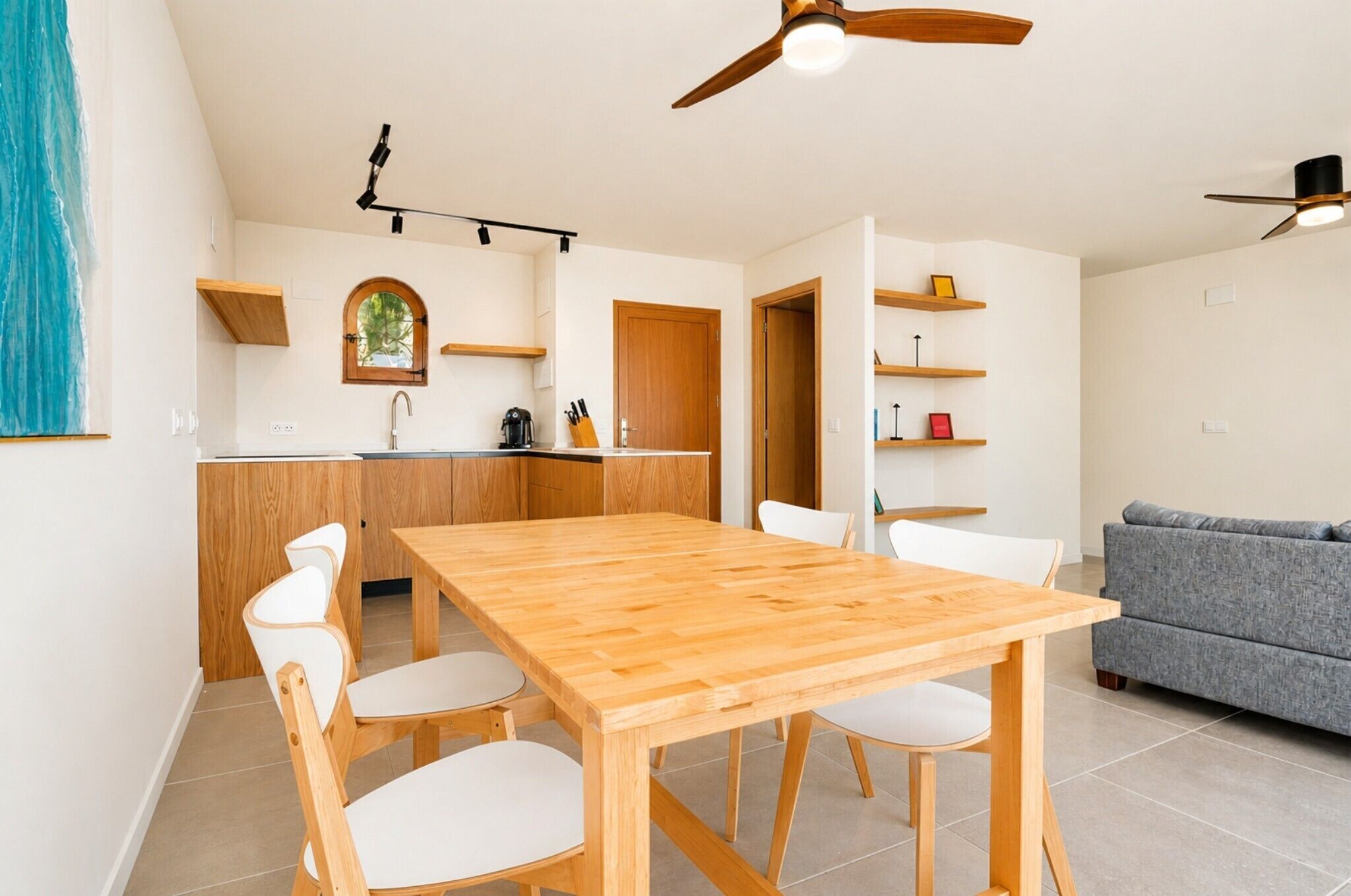 A modern kitchen with wooden cabinets, a dining table with chairs, a grey sofa, and a ceiling fan.