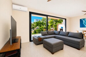 A modern living room with a sectional sofa, ottoman, flat-screen TV, and large windows offering a view of buildings and greenery. - Holiday Home 'Mía' with Sea View, Wi-Fi and Air Conditioning (Xàbia)