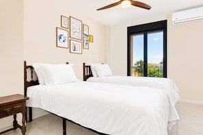 A hotel room with two beds, a wooden nightstand, a ceiling fan, and a large window with a view of greenery.