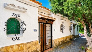 Una estrecha calle empedrada con un edificio blanco que presenta una base de piedra, un ventanal verde y una verja negra.