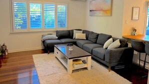Sala de estar com um sofá cinza, uma mesa de centro, um tapete branco e uma janela com persianas.