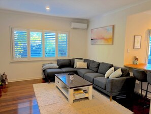 Ein Wohnzimmer mit grauem Sofa, Couchtisch, weißem Teppich und einem Fenster mit Jalousien.
