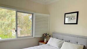 Um quarto com uma cama, uma janela com persianas, uma mesa de cabeceira com um vaso e um quadro na parede.