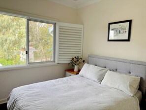 Ein Schlafzimmer mit Bett, Fenster mit Jalousien, Nachttisch mit einer Vase und einem gerahmten Bild an der Wand.