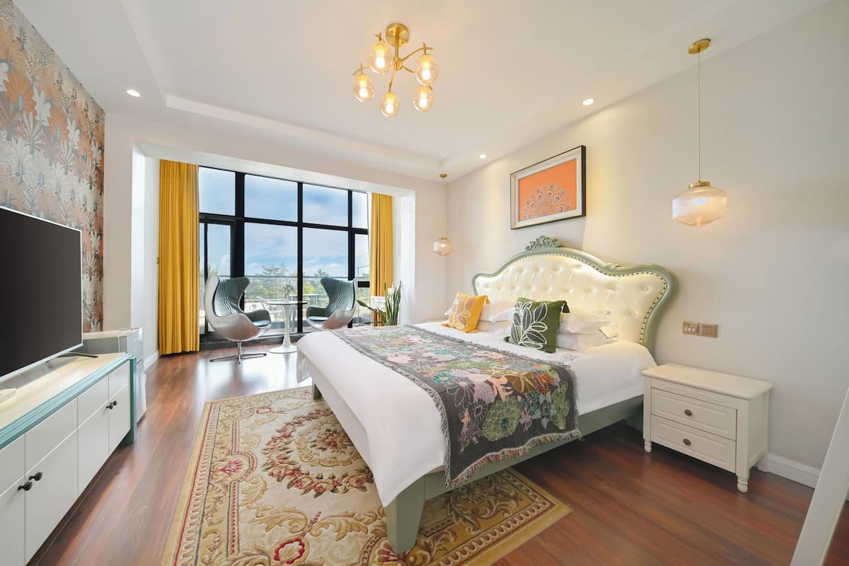 A bedroom with a large bed, a TV, a bedside table, a nightstand, a chandelier, and a view of the outdoors.