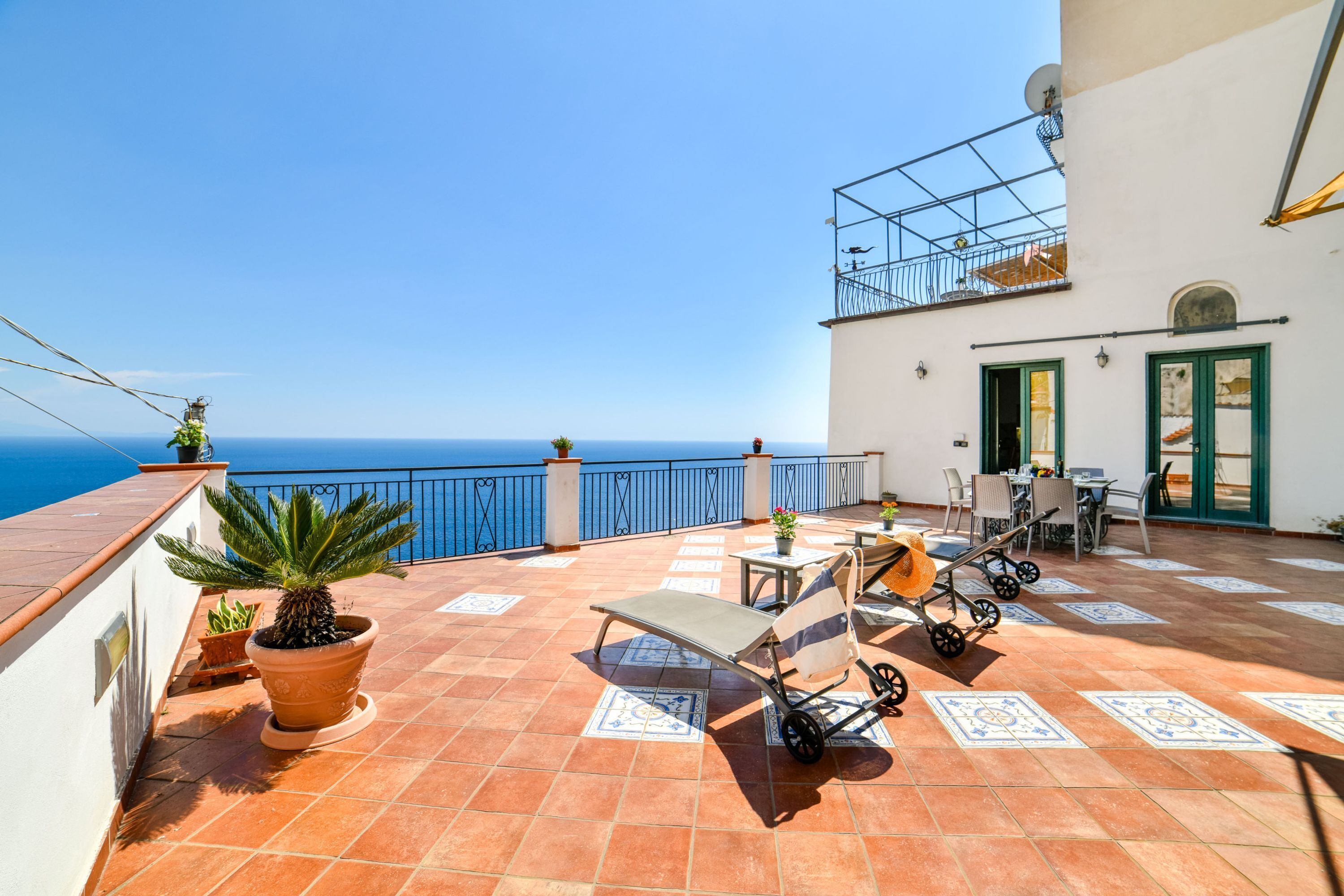 A terrace with a view of the sea, a table with chairs, a sun lounger, and potted plants.