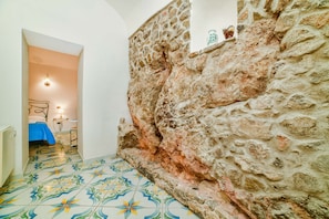 Ein schmaler Flur mit einer Steinwand, einem Bett mit blauer Bettdecke und einem gefliesten Boden mit floralem Muster.
