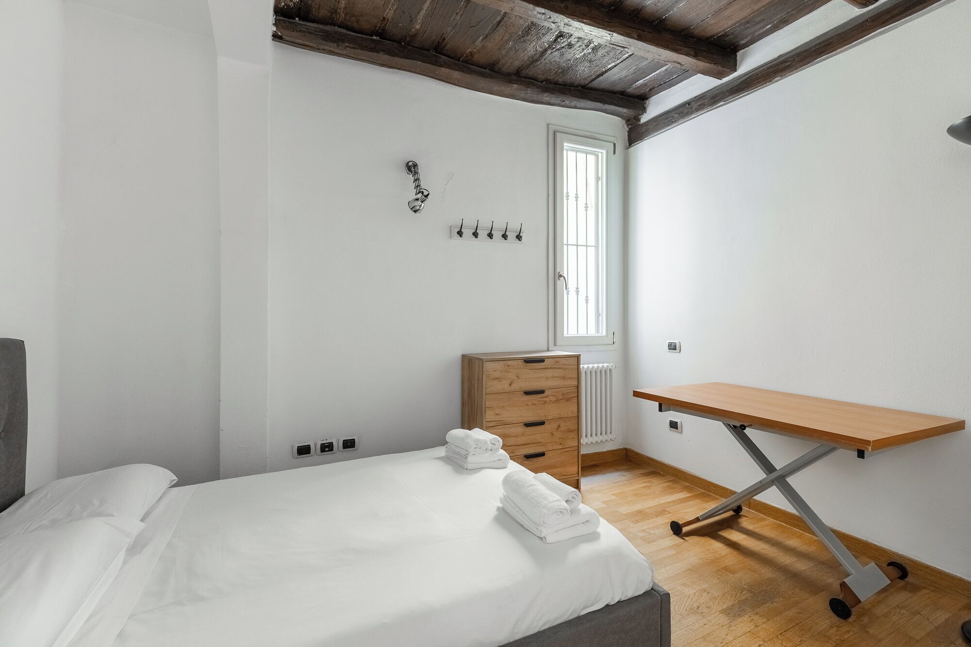 A bedroom with a bed, a wooden dresser, a desk, a radiator, and a window with a view.