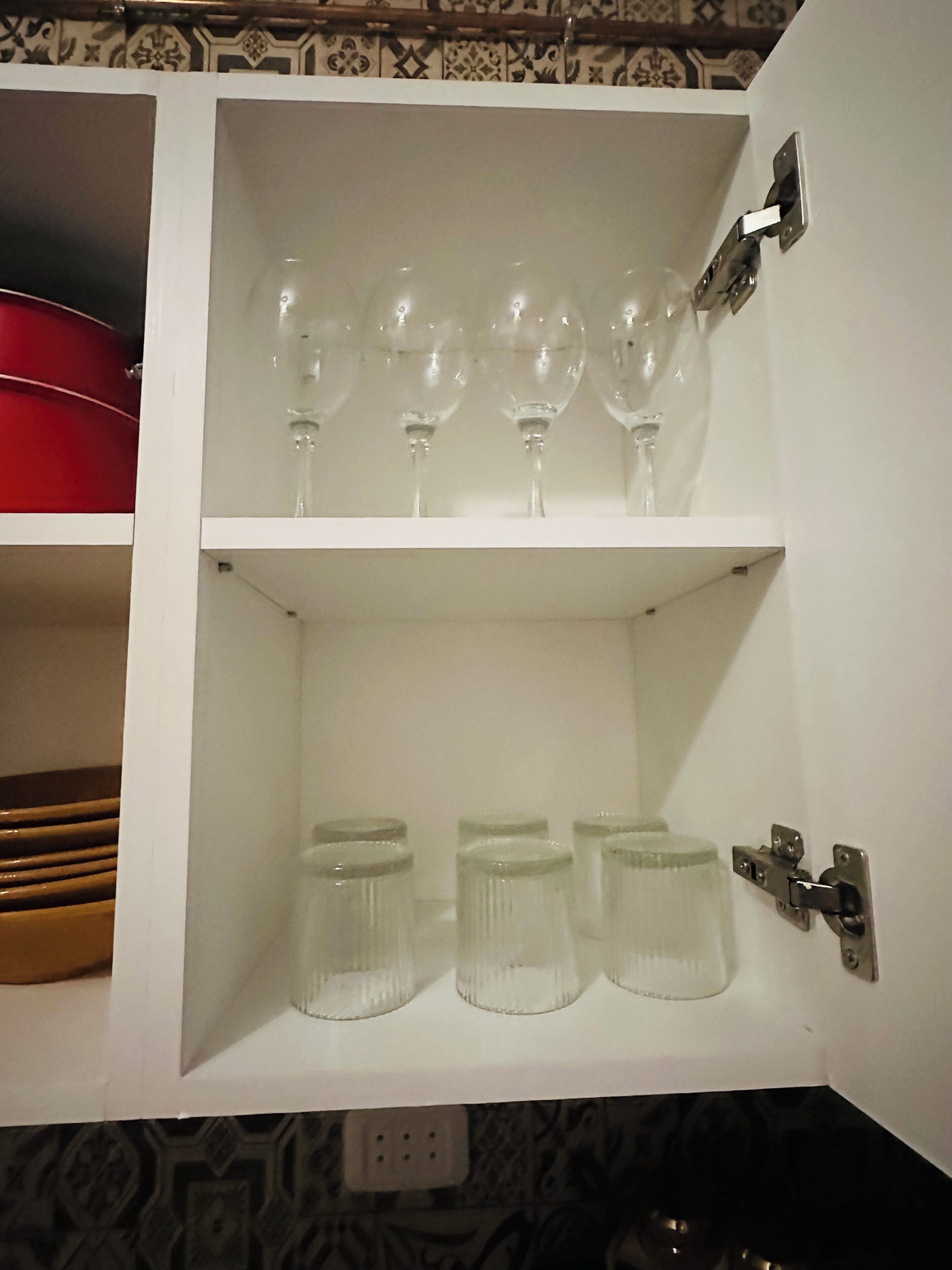 Un mueble blanco con copas de vino en la repisa superior y frascos de vidrio en la inferior.