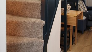 A carpeted staircase with a glass handrail, leading to a room with a wooden table and a dark-colored sofa. - The Sails – Romantic Retreat for Two, Cosy Light-Filled House (Leek)