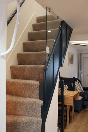 A carpeted staircase with a glass handrail, leading to a room with a wooden table and a dark-colored sofa. - The Sails – Romantic Retreat for Two, Cosy Light-Filled House (Leek)