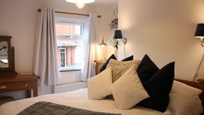 A bedroom with a bed, a bedside table, a lamp, a window with curtains, and a wall-mounted light fixture. - The Sails – Romantic Retreat for Two, Cosy Light-Filled House (Leek)