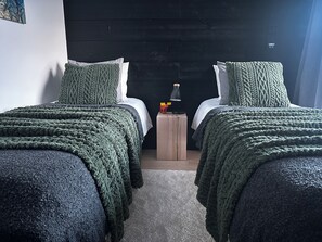 Ein Zimmer mit zwei Betten, jeweils mit einer grünen, gestrickten Decke, einem kleinen Nachttisch mit Lampe und einer wandmontierten Leuchte.