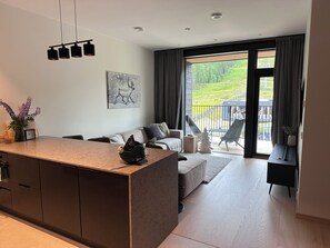 Eine moderne Küche mit einer zentralen Kochinsel, ein Essbereich mit einer Couch und Stühlen sowie ein Balkon mit Blick auf Grünflächen.