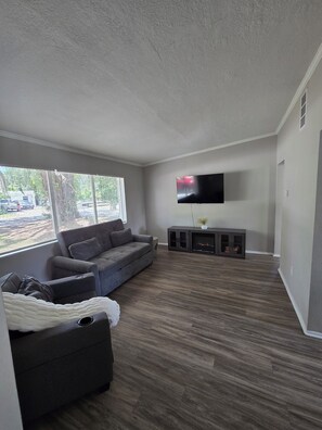 Ein Wohnzimmer mit grauem Sofa, einem an der Wand befestigten Flachbildfernseher und Holzboden.