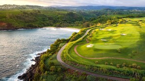 A coastal golf course with a winding path, a sandy beach, and a view of distant hills.