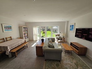 Ein Wohnzimmer mit einer Couch, einer Bank, einem Bücherregal, einem Fernseher und einer Glastür zum Garten.