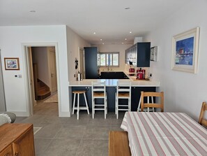 Et moderne køkken med en central ø, hvide barstole og et spiseområde med dug med striber.