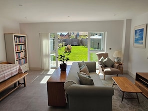 Ein Wohnzimmer mit einer Couch, einem Holztisch, einem Bücherregal und Blick auf einen grünen Garten durch eine Glastür.
